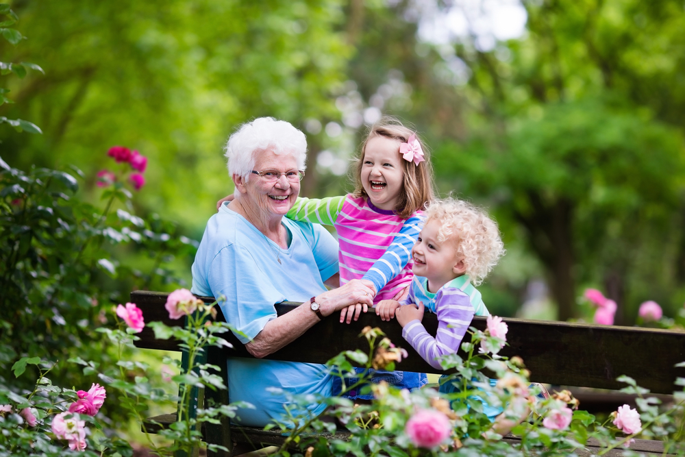Happy senior lady playing with little boy and girl in blooming rose garden. Grandmother with grand children sitting on a bench in summer park with beautiful flowers. Kids gardening with grandparent.